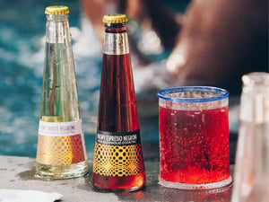Negroni mocktail bottles and a chilled red drink sit poolside on a sunny day.