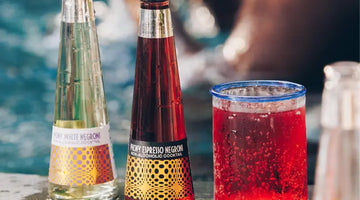 Negroni mocktail bottles and a chilled red drink sit poolside on a sunny day.