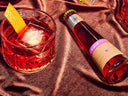 Non-alcoholic Negroni shown in a glass with an orange peel beside a vibrant bottled mocktail on satin fabric