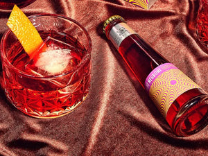 Non-alcoholic Negroni shown in a glass with an orange peel beside a vibrant bottled mocktail on satin fabric