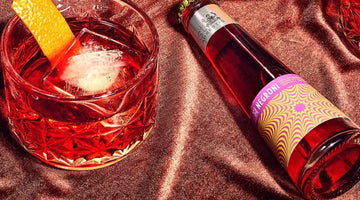 Non-alcoholic Negroni shown in a glass with an orange peel beside a vibrant bottled mocktail on satin fabric