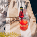 Alcohol free negroni in a glass with ice beside a bottled Phony Negroni on a striped towel in dappled sunlight.