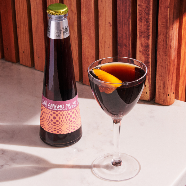 Virgin Amaro Falso non-alcoholic cocktail in a stemmed glass with an orange twist beside its bottled version on a marble surface.